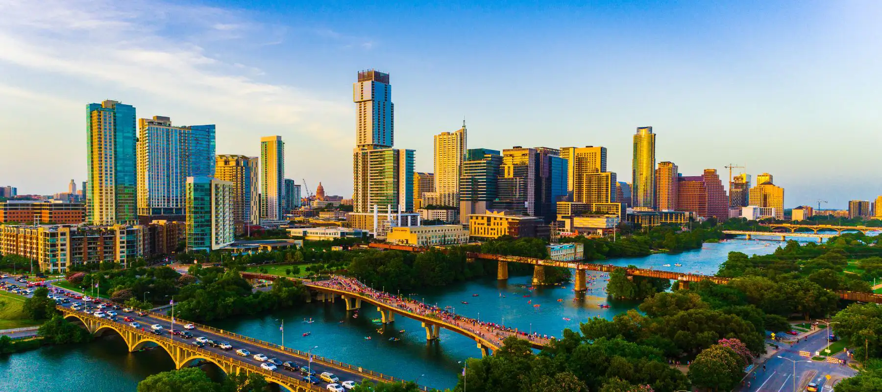 A vibrant city skyline with modern high-rise buildings, a river running through the city, multiple bridges, lush greenery, and people enjoying outdoor activities under a clear blue sky at sunset.