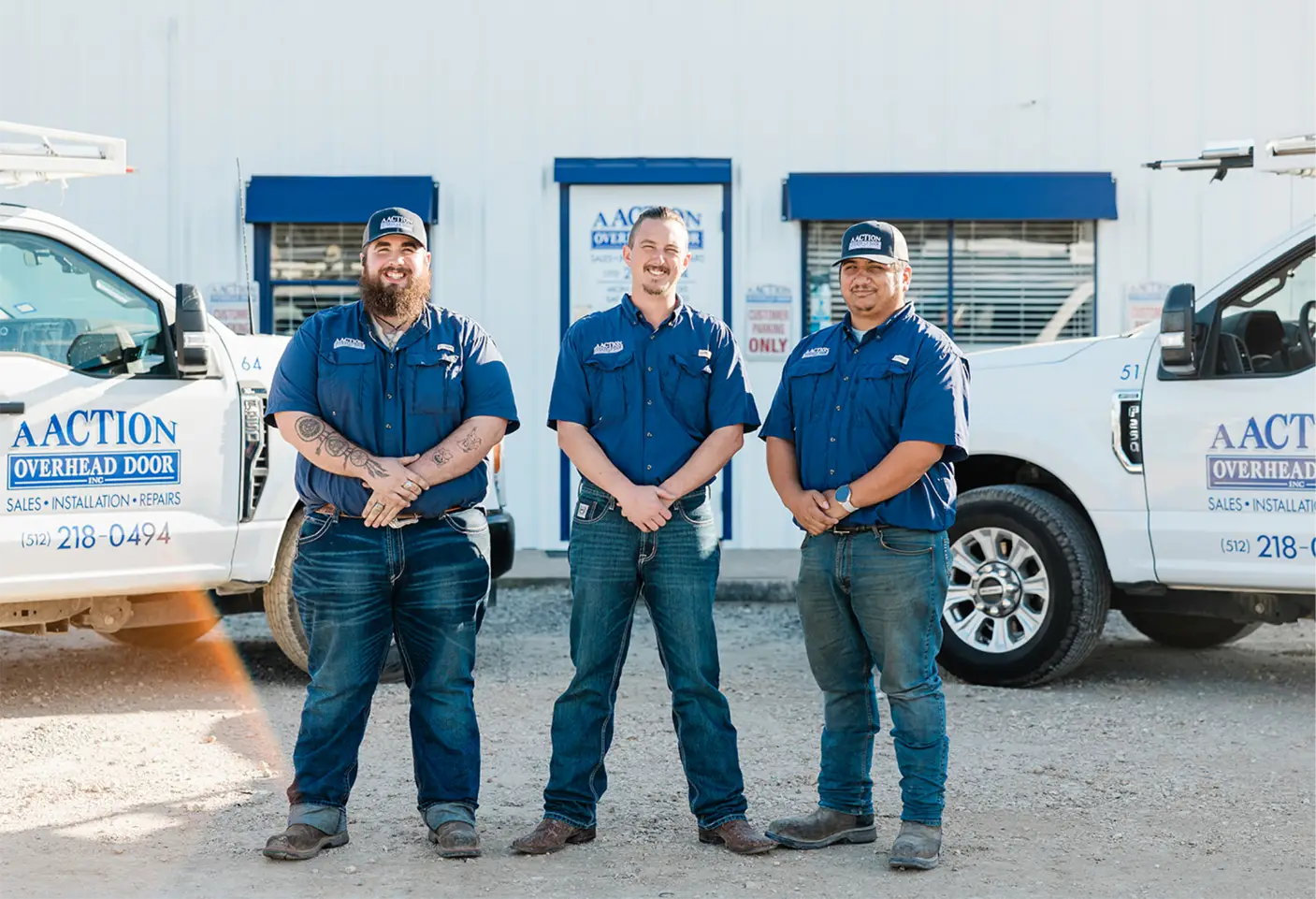 Three men wearing blue shirts and jeans stand smiling in front of a white building, flanked by two white work trucks with AACTION Overhead Door logos.