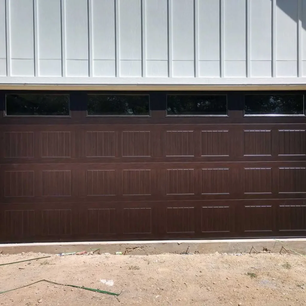A closed, dark brown garage door with rectangular panels and three narrow horizontal windows at the top, set in a white paneled exterior wall. The ground in front is bare dirt.