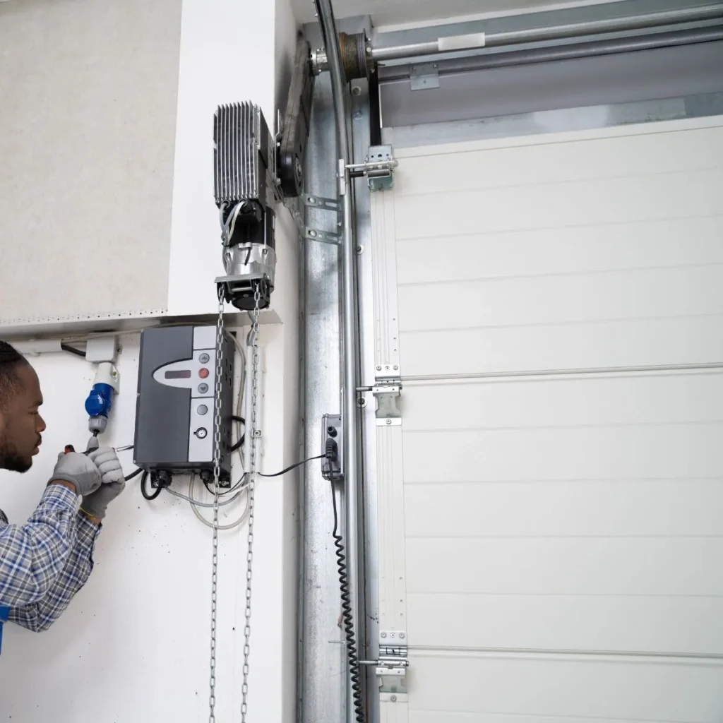 A person wearing gloves installs or repairs a control box connected to an automatic garage door opener inside a garage. The person is using a tool to work on the electrical components.