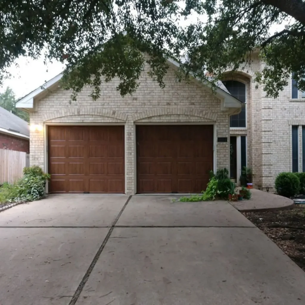 A suburban house with a light brick exterior, two large brown garage doors, a concrete driveway, and some green plants near the entrance under a tree canopy.
