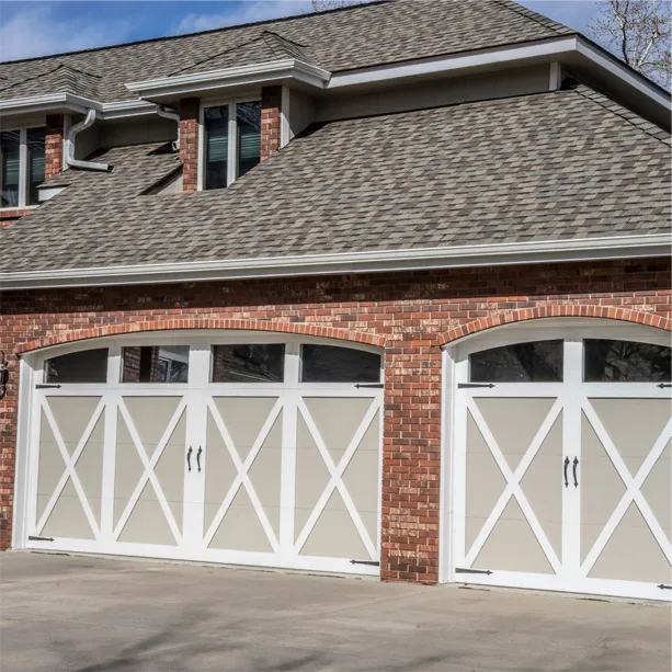 A brick house with three white garage doors featuring a crisscross pattern and black handles. The house has gray shingles, double windows above the garage, and a clean, paved driveway.