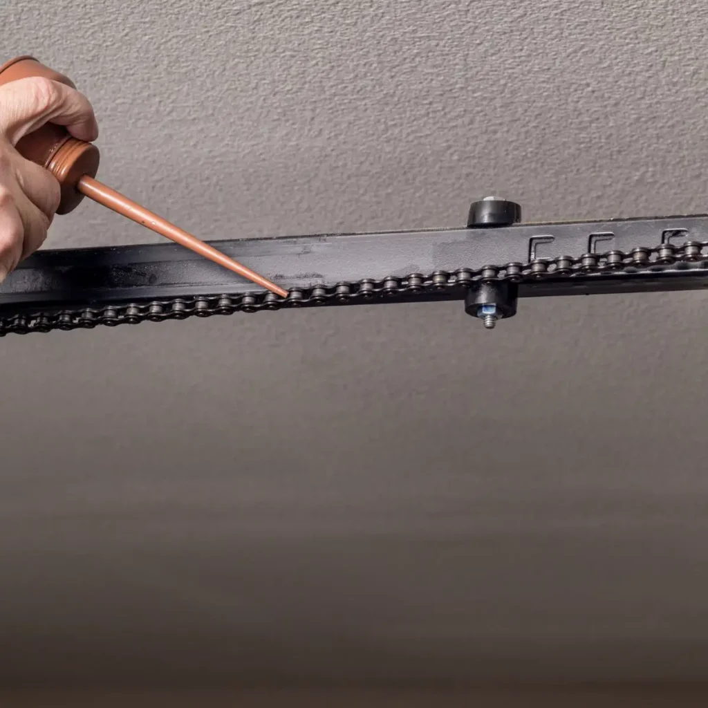 A hand is applying oil to a metal chain on a garage door opener rail mounted to the ceiling.