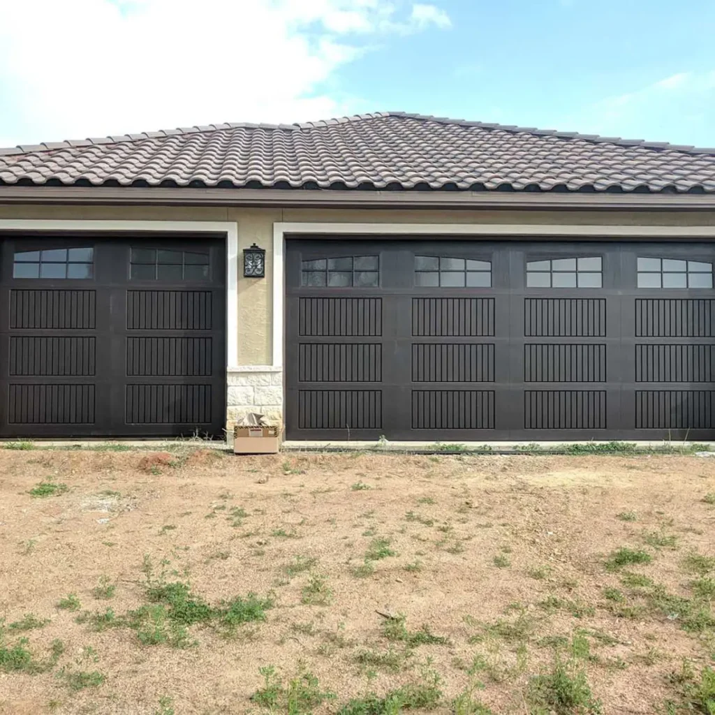 Two black garage doors with vertical and square panel designs on a beige house with a tiled roof. There is a small box placed on the ground in front of the house, and the yard is mostly bare dirt with sparse grass.