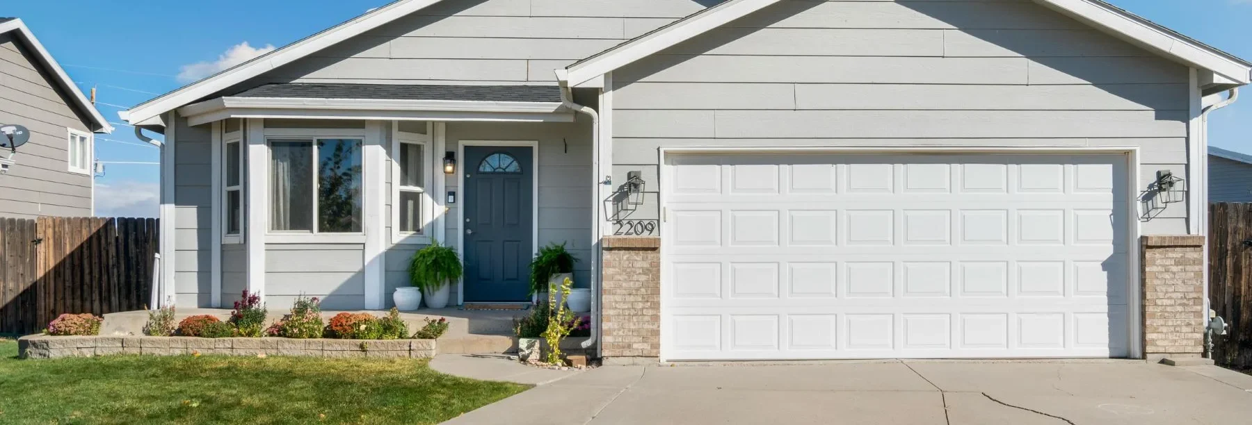 Single-story house with light gray siding, a dark gray front door, and a white double garage. There’s a small front porch with potted plants, flowers, and a manicured lawn bordered by a low retaining wall.