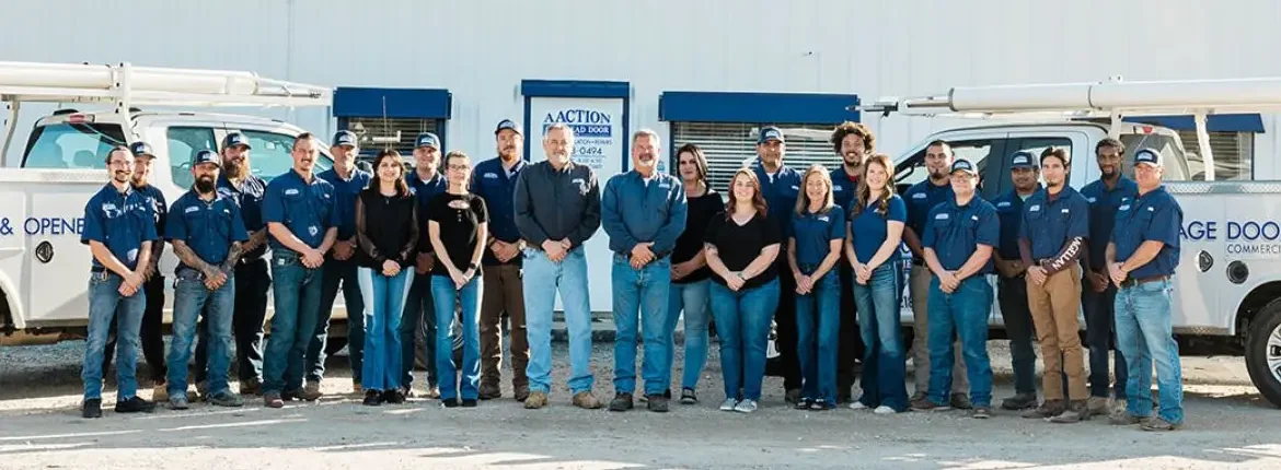 A group of 22 people, wearing blue shirts and jeans, stand in front of a white building with blue trim and two white work trucks. They appear to be a team, posing for a company photo outdoors.