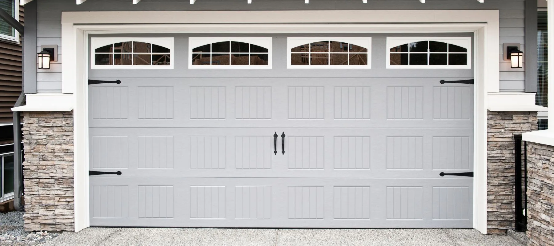 A modern gray garage door with vertical panels, decorative black handles, and hinges. The top of the door has four arched windows with grid patterns. The garage is framed by stone and white trim.