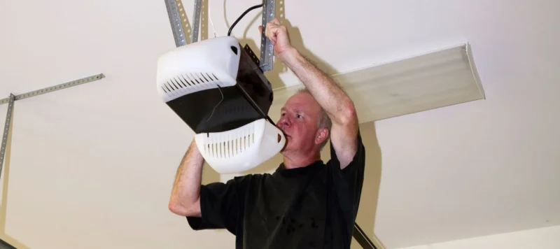 A man standing on a ladder installs or repairs a garage door opener mounted to the ceiling, using a tool to adjust or secure the device.