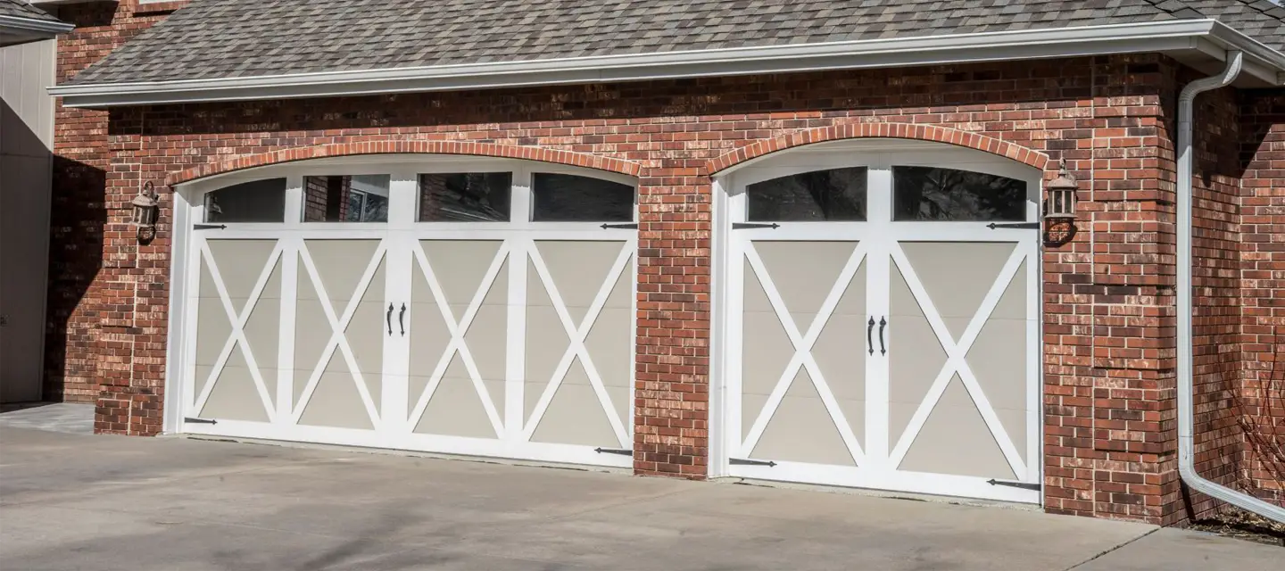 Two white garage doors with decorative X-shaped trim and four upper window panes each, set in a red brick wall of a house with a shingled roof. There are outdoor lanterns mounted next to each door.