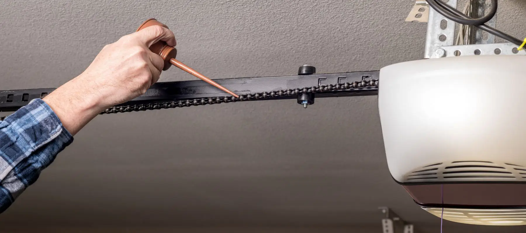 A person applies lubricant to the chain of a garage door opener mounted on the ceiling, using a small oil can.