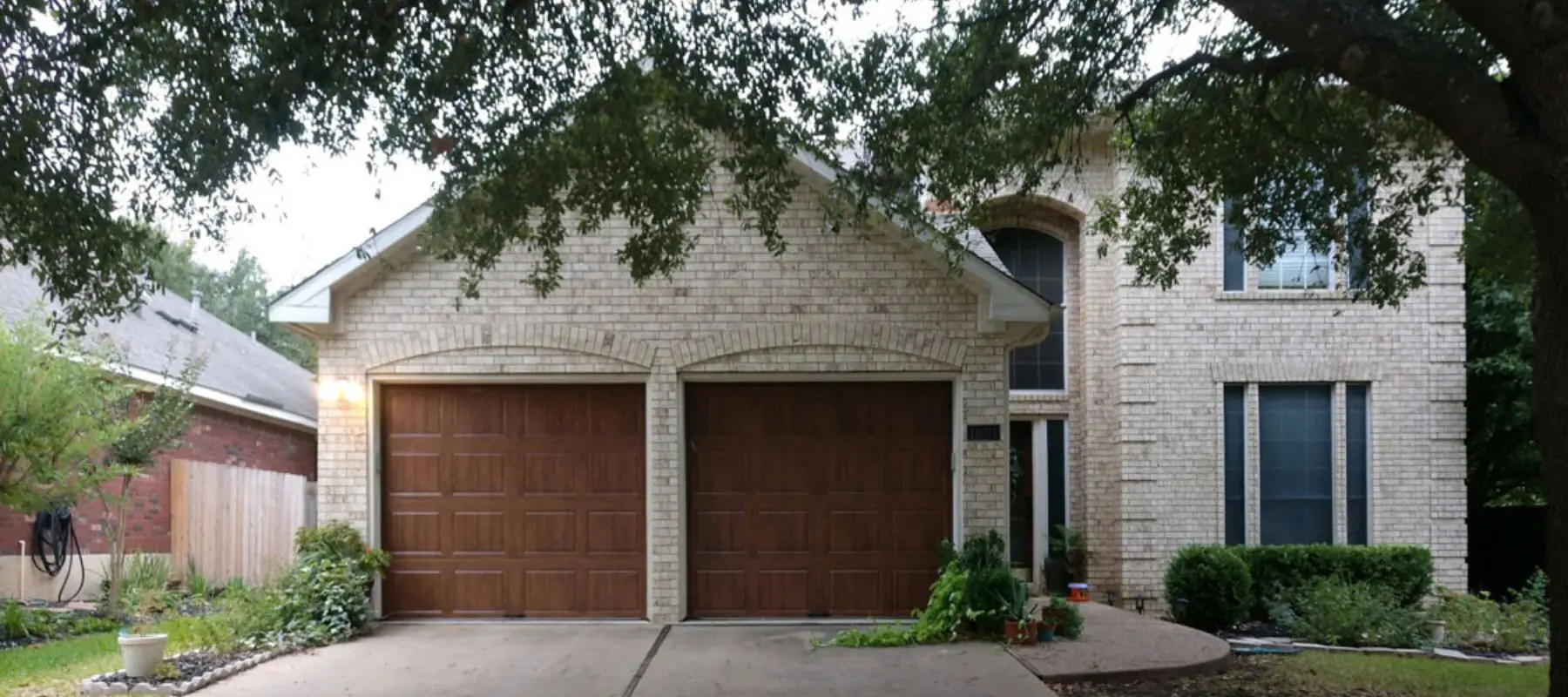 A two-story brick house with two wooden garage doors, an arched entranceway, tall windows, and a shaded driveway bordered by plants and trees.