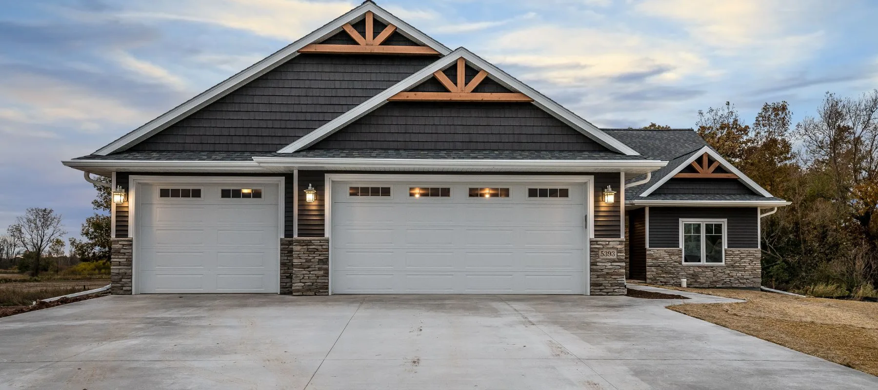 A modern single-story house with dark gray siding, stone accents, and a large three-car white garage. The driveway is wide and clean, and there are trees and grass surrounding the home.