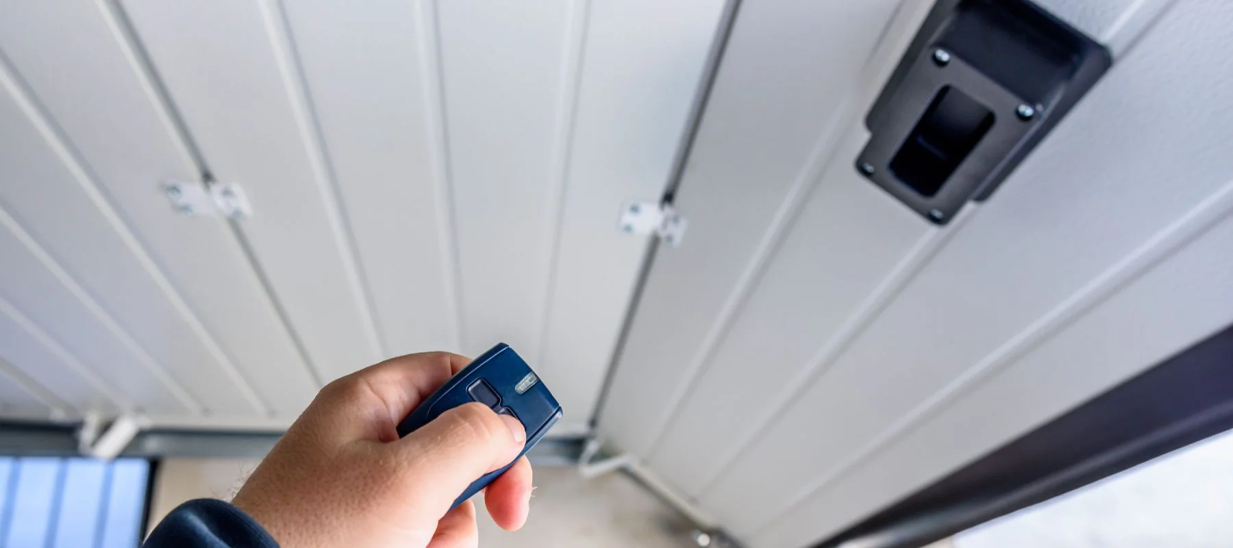 A person’s hand holding a remote control points upward toward a closed garage door, with a black garage door opener mechanism visible on the ceiling.