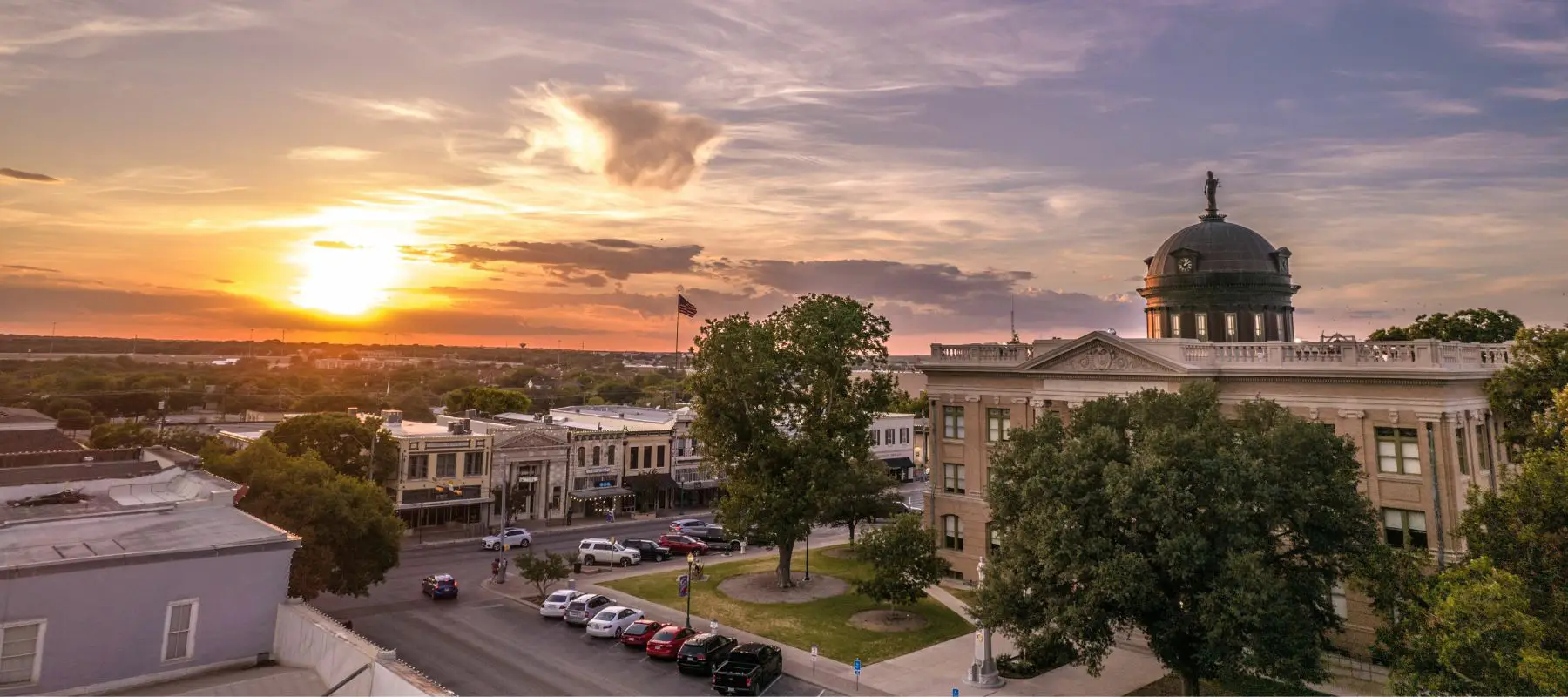 A picturesque sunset over a small town square, featuring historic buildings, a domed courthouse, trees, and parked cars, with a vibrant sky filled with clouds and sunlight.