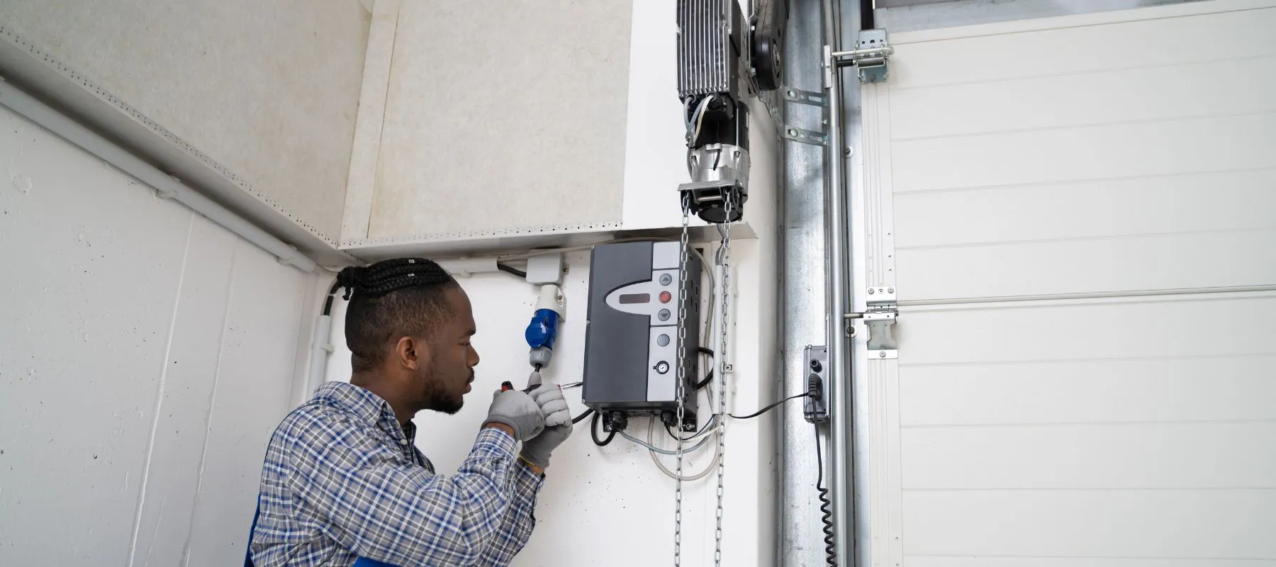 A technician wearing gloves and a plaid shirt inspects or repairs an electronic control box mounted on a wall near a garage door mechanism.