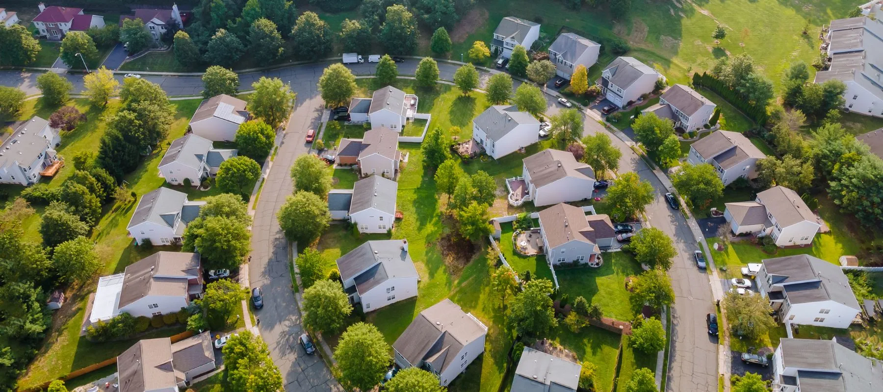 Aerial view of a suburban neighborhood with curved streets, single-family houses, driveways, and green lawns, surrounded by trees and greenery on a sunny day.