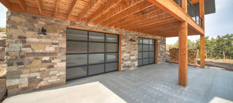 View of a modern house exterior with a stone facade, two large glass garage doors, exposed wooden beams, and a stack of firewood beside the building, with trees visible in the background.