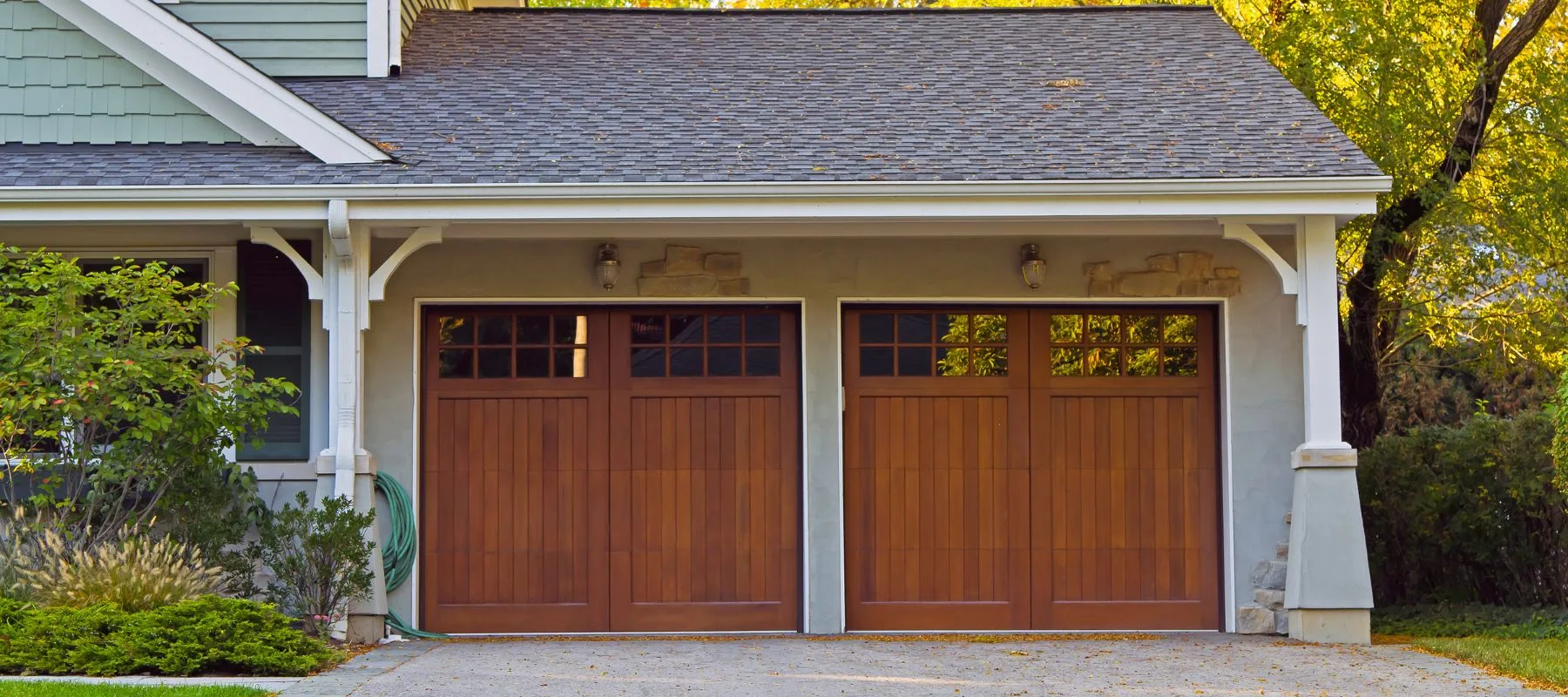 A suburban house features a double garage with two closed wooden doors, each with small square windows at the top. The driveway is empty, and greenery surrounds the building.
