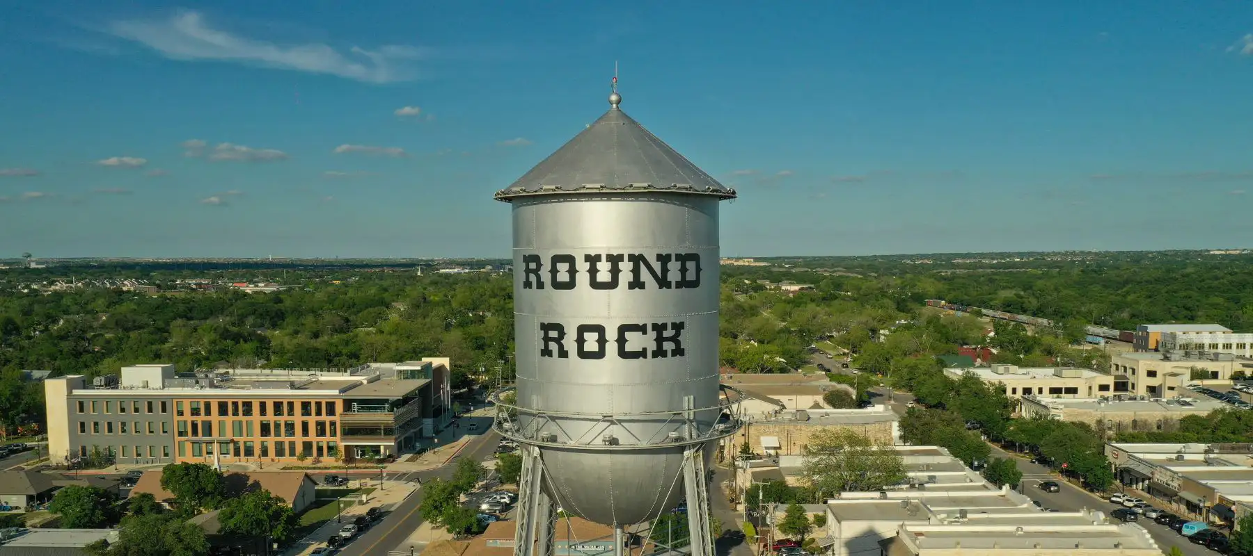 A silver water tower with the words Round Rock stands prominently over a small city with buildings, roads, and green trees under a clear blue sky.