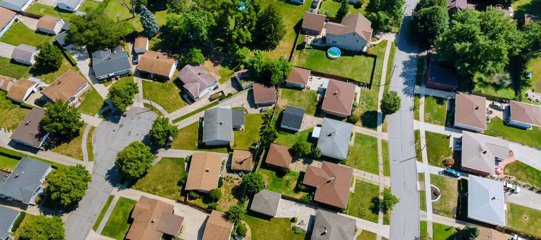 Aerial view of a suburban neighborhood with houses, green lawns, trees, driveways, and a circular street, taken on a sunny day. Some yards have pools and outdoor furniture.