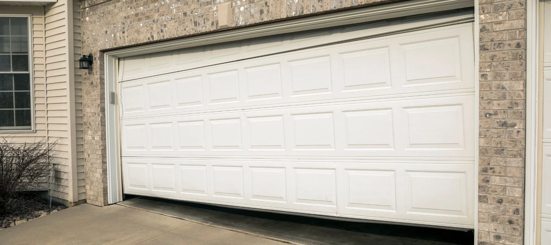 A white garage door is stuck halfway open, tilting unevenly on its tracks against a brick and siding exterior wall, indicating it may be broken or off-track.