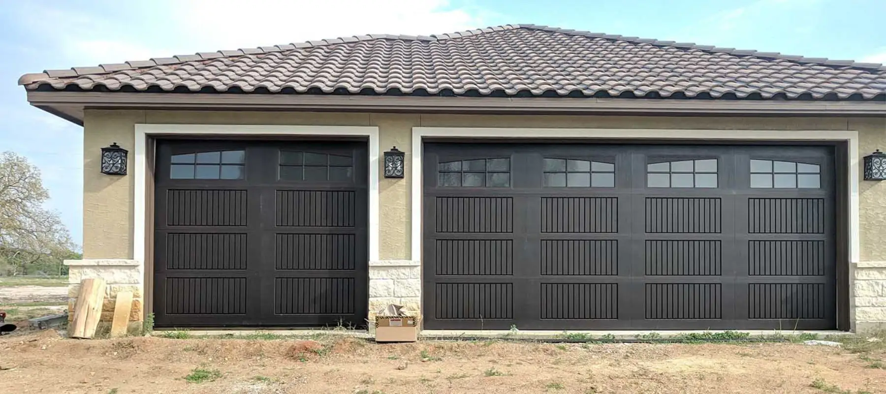 Two large, dark-colored garage doors with rectangular panels on a light-colored house with a tiled roof. A small package sits on the ground between the doors, with some bare soil and grass in front.