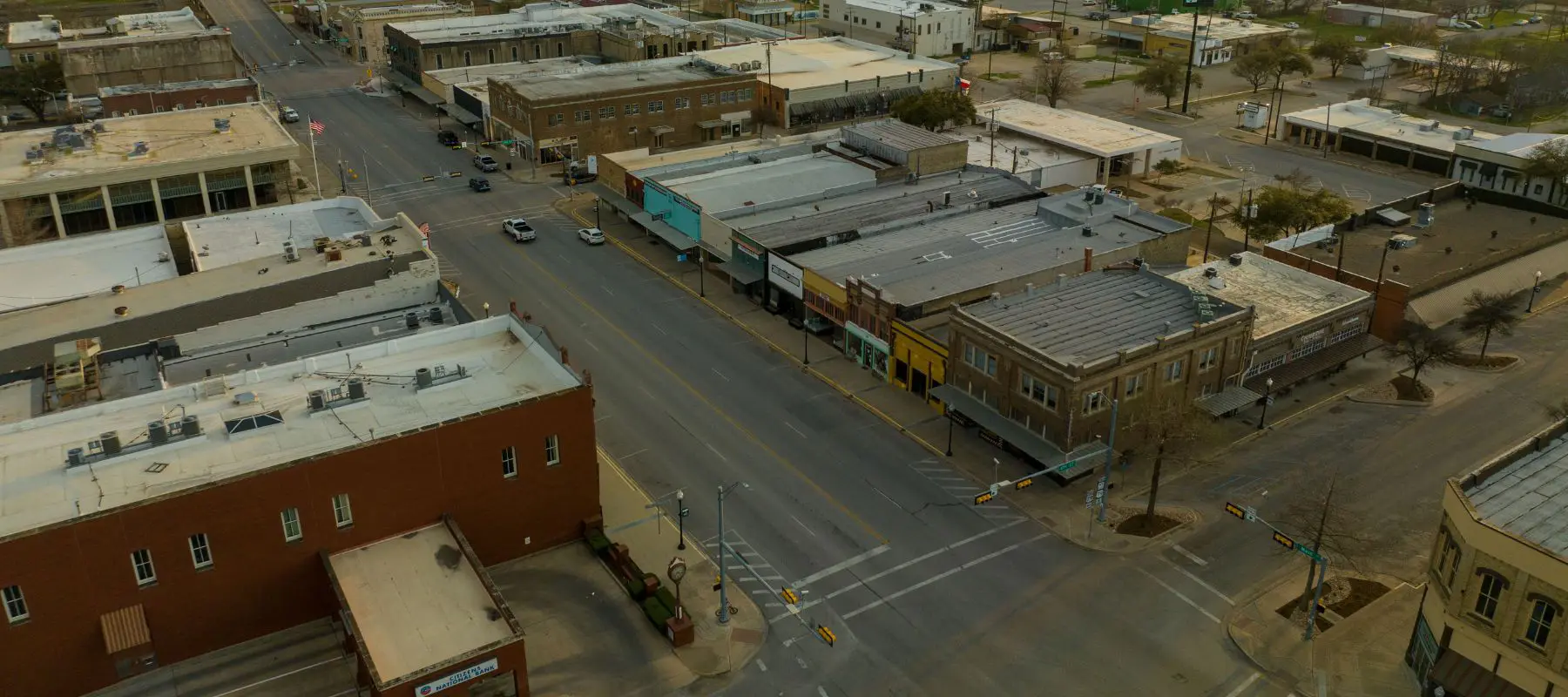 Aerial view of a quiet small town intersection with empty streets, low-rise buildings, and shops lining both sides of the road on a cloudy day.