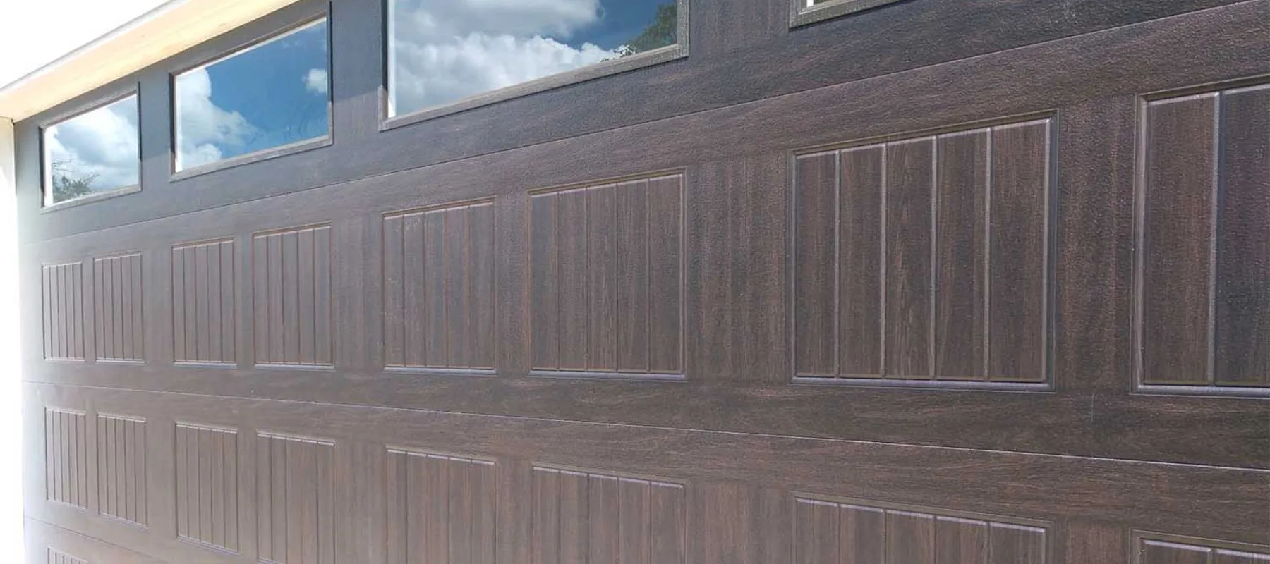 Close-up view of a dark brown, paneled garage door with rectangular windows at the top reflecting a partly cloudy sky.