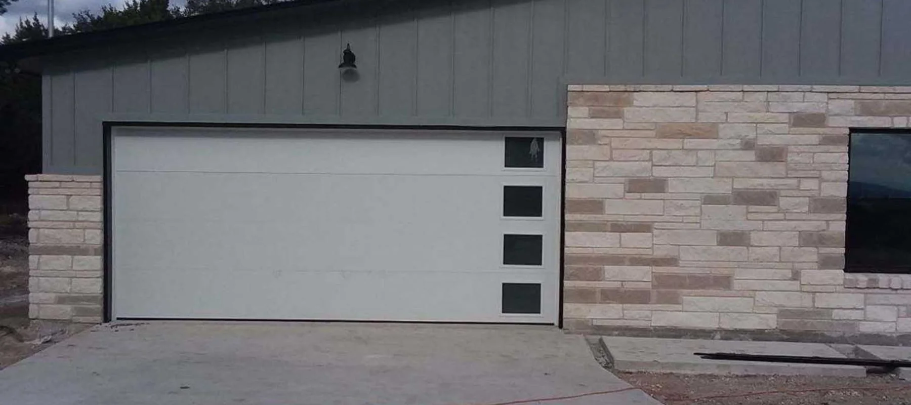 A modern house exterior with a gray garage door featuring four square windows arranged vertically on the right side, next to a wall with light-colored stone brickwork.