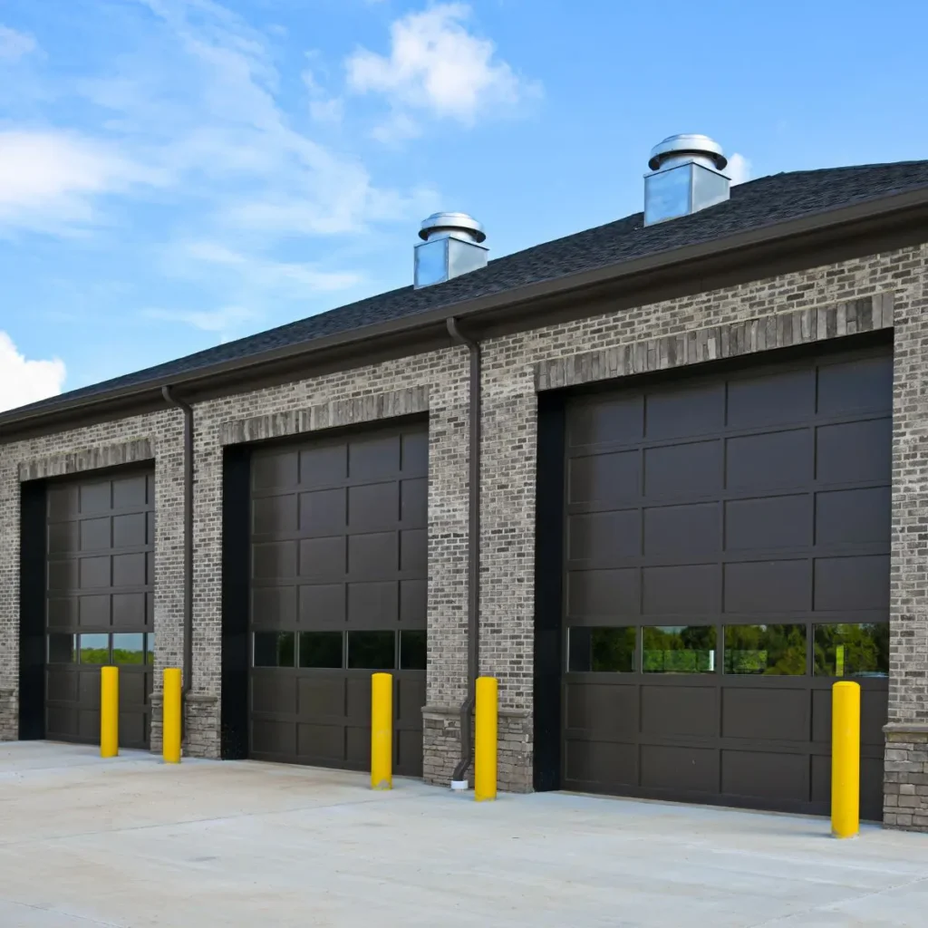 A modern brick building with three large dark brown garage doors, each with windows, and yellow safety bollards in front. The sky is blue with some clouds.