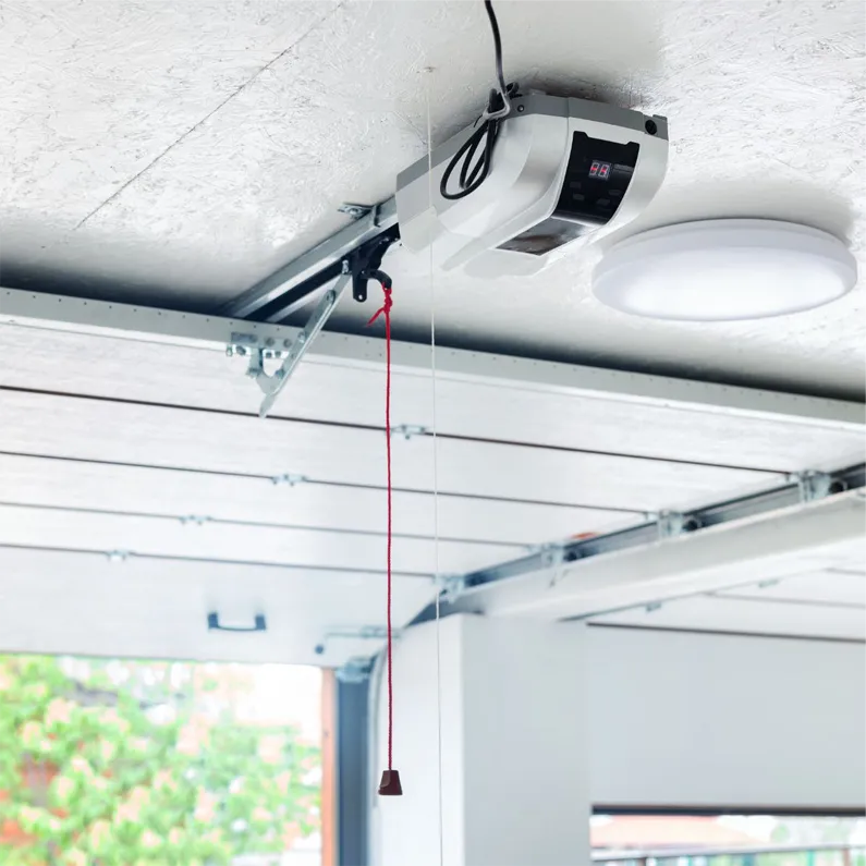 A garage door opener mounted on the ceiling with a red emergency release cord hanging down and part of an open garage door visible below.