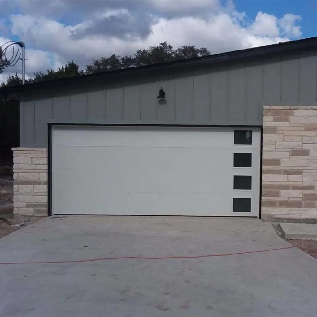 A garage door with four small square windows arranged vertically on the right side. The building has gray siding on top and light stonework on the lower portion, with a cloudy sky in the background.