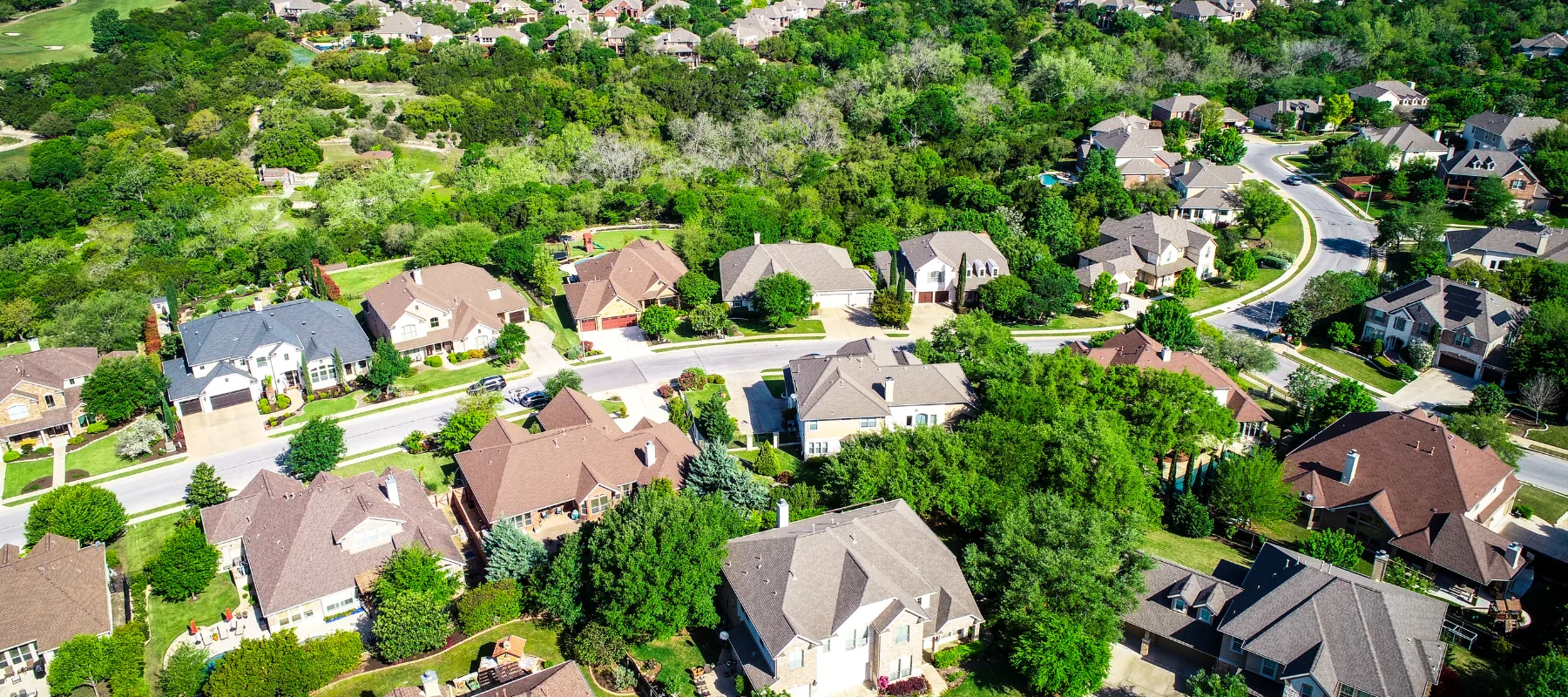 Aerial view of a suburban neighborhood with tree-lined streets, large single-family homes, green lawns, and curving roads surrounded by dense forest and greenery.