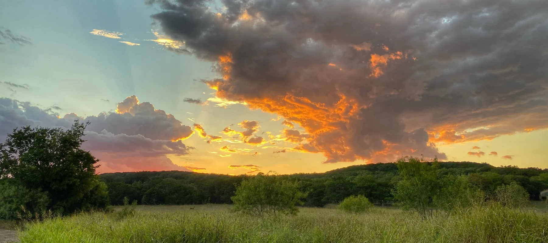 A dramatic sunset sky with vibrant orange and yellow clouds, partially obscured by dark gray clouds, above a green field and tree-covered hills.