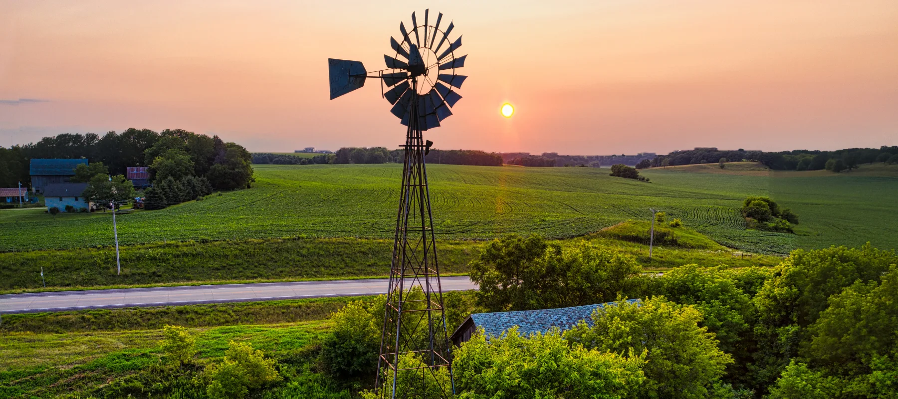 A windmill stands tall in front of a lush green field at sunset, with the sun low in the sky and a country road nearby, surrounded by trees and distant farm buildings.