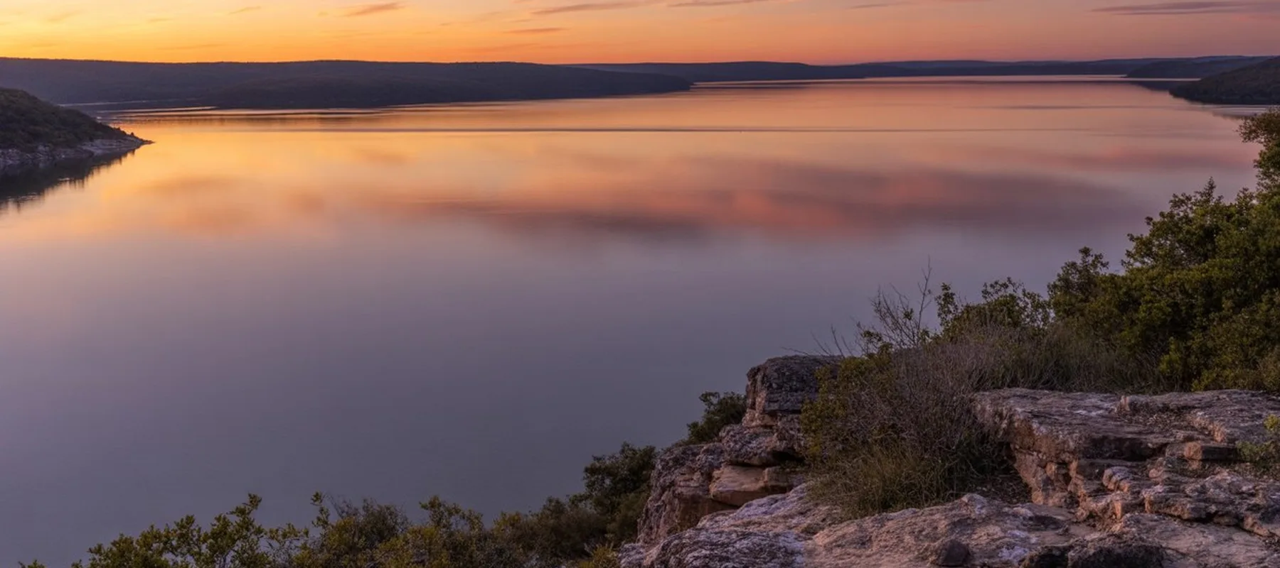A calm lake reflects the orange and purple hues of a sunset sky, bordered by distant hills and rocky cliffs with sparse greenery in the foreground.