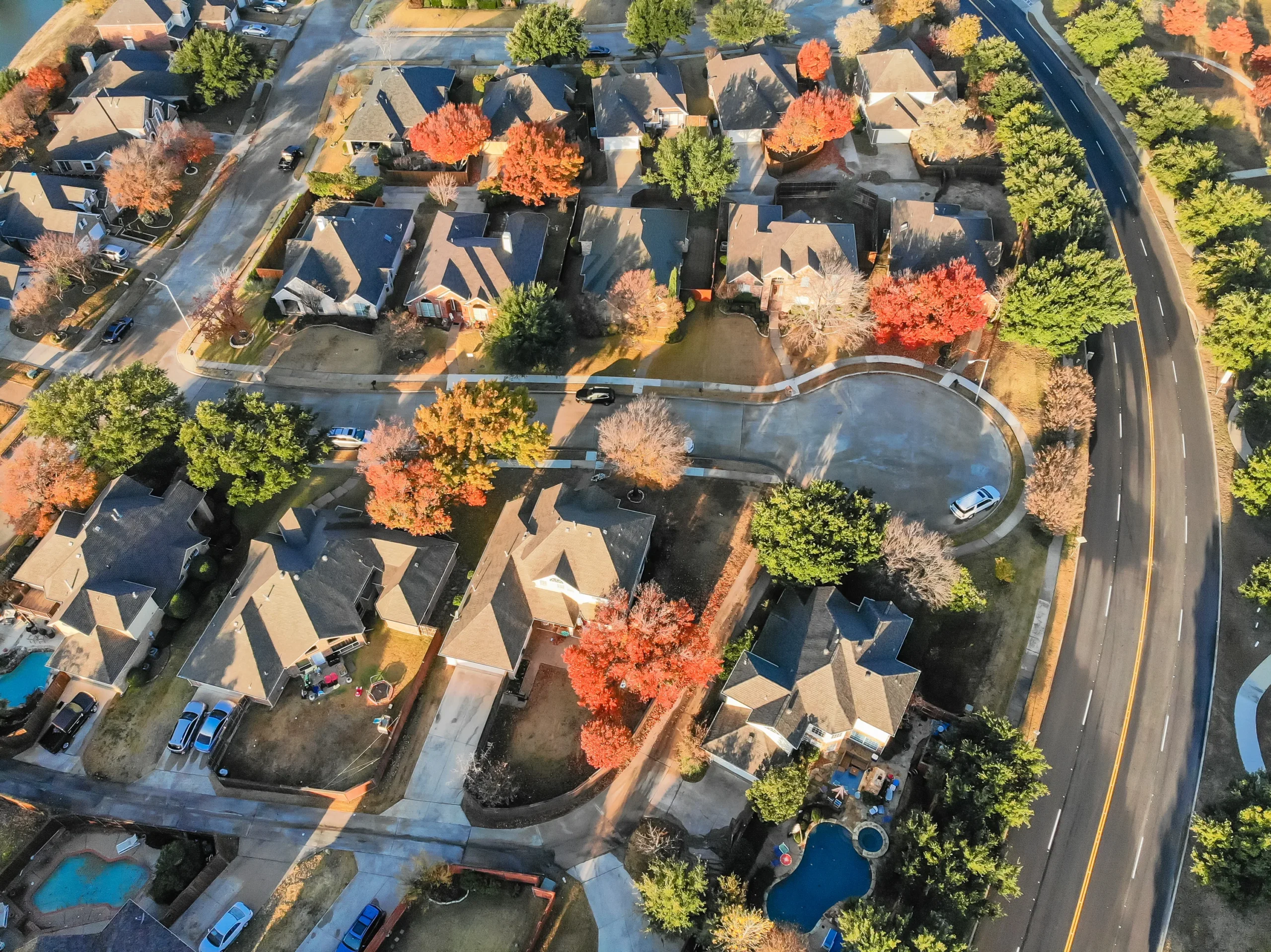 Aerial view of a suburban neighborhood with houses, driveways, and streets. Some trees, vibrant with autumn colors, are scattered among green ones. A curved road borders the area, and pools are visible in backyards.