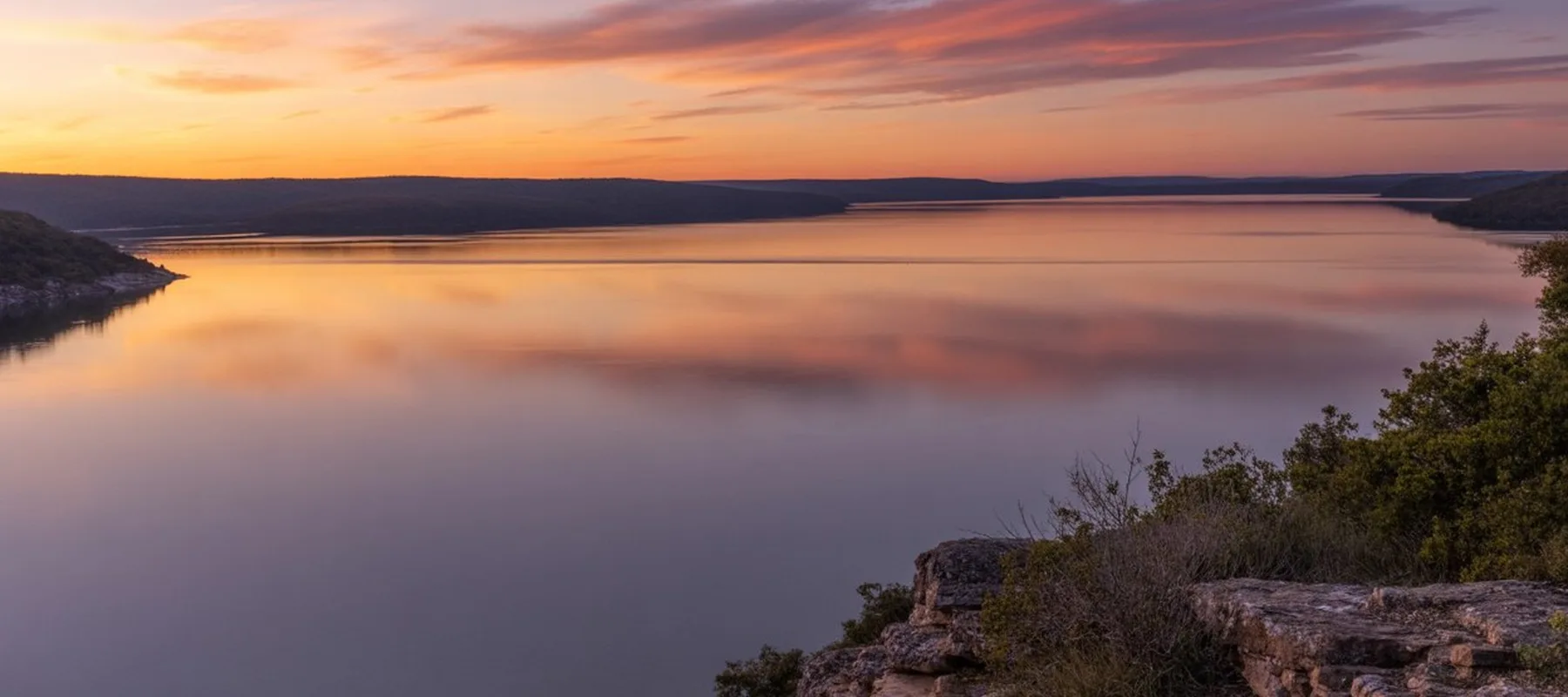 A calm lake stretches into the distance, reflecting a colorful sunset sky with orange and pink clouds. Hills and trees line the shore, and rocky terrain is visible in the foreground.