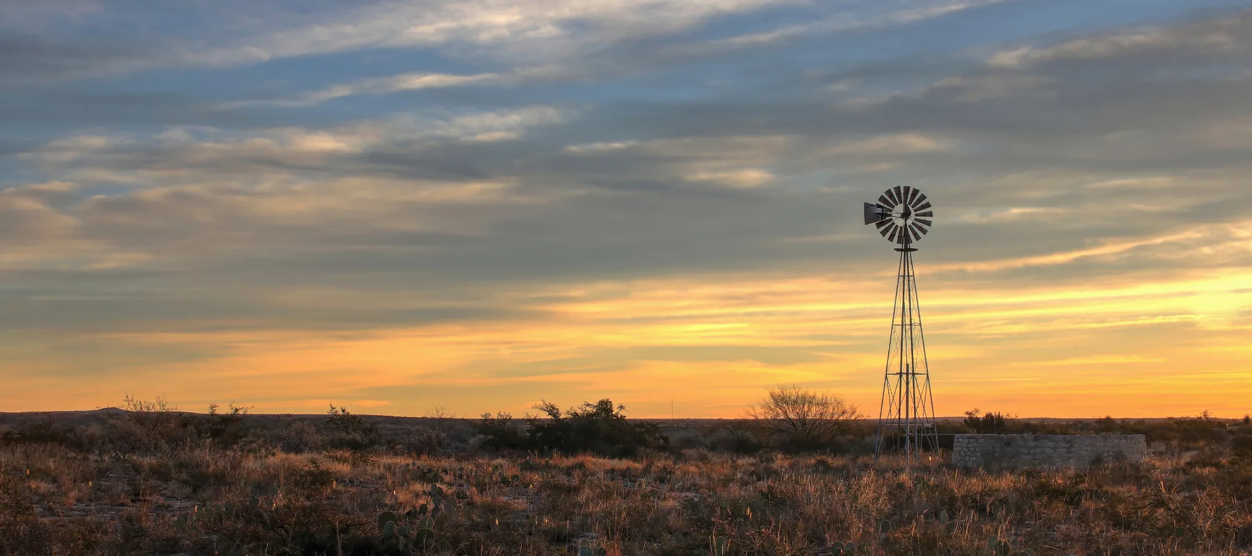 A windmill stands in an open field at sunset, with golden light illuminating dry grasses and low bushes under a partly cloudy sky.